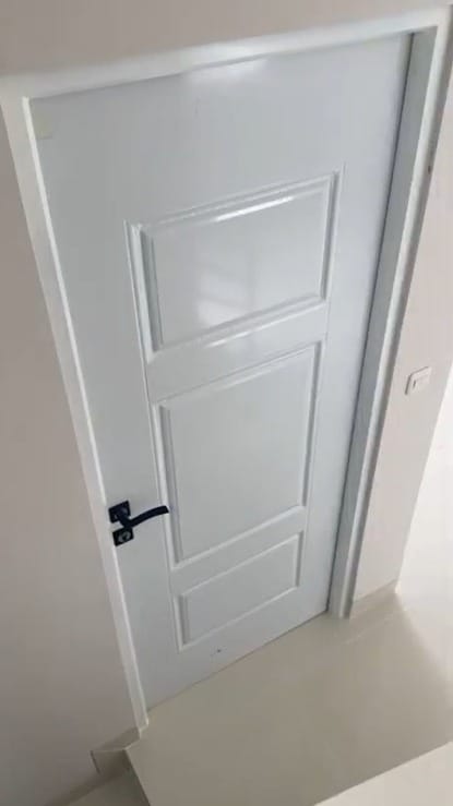 Pintu interior minimalis berwarna putih dengan gagang pintu hitam modern, menampilkan desain tiga panel persegi panjang yang ditinggikan, berdiri di atas lantai ubin berwarna krem di lorong yang cerah.